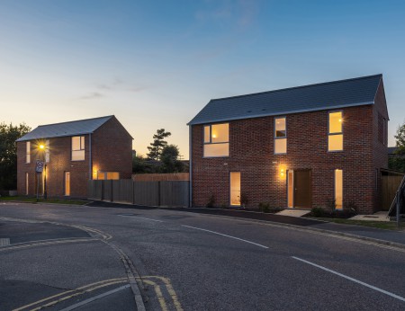 An architectural CGI illustration of two red brick modern cottages at dusk. Internal lights are on. The daylight is fading.